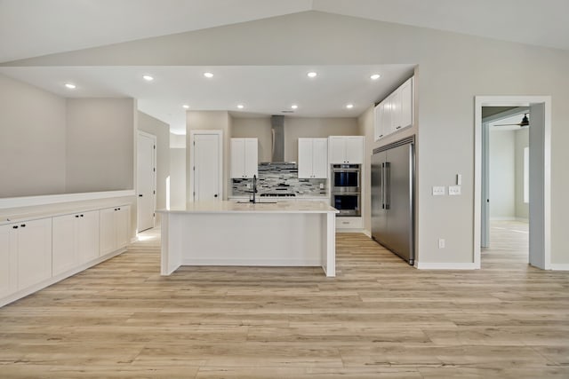 kitchen with appliances with stainless steel finishes, a kitchen island with sink, wall chimney exhaust hood, and vaulted ceiling