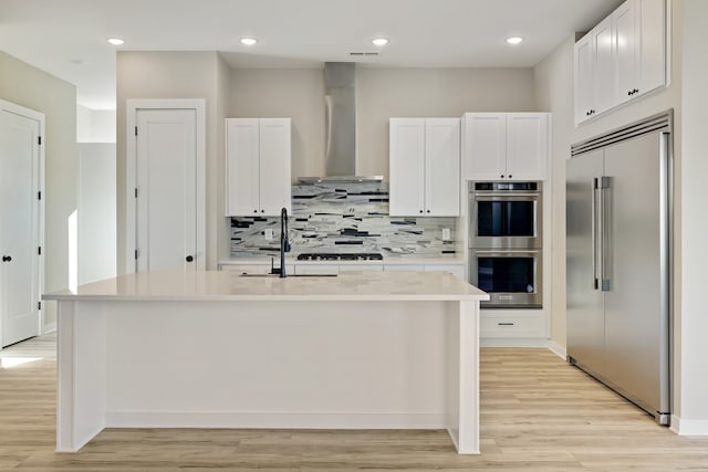 kitchen featuring a kitchen island with sink, sink, white cabinetry, wall chimney range hood, and appliances with stainless steel finishes