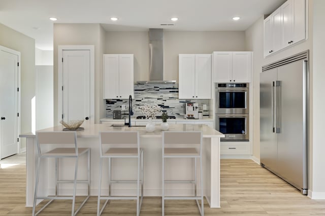 kitchen with an island with sink, wall chimney range hood, white cabinetry, stainless steel appliances, and a kitchen bar