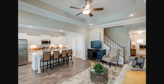 living room with a tray ceiling, ceiling fan, ornamental molding, and light wood-type flooring