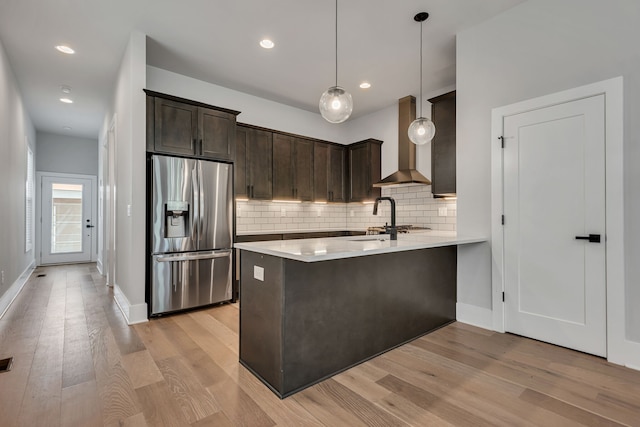 kitchen with light wood-type flooring, kitchen peninsula, wall chimney exhaust hood, decorative light fixtures, and stainless steel fridge