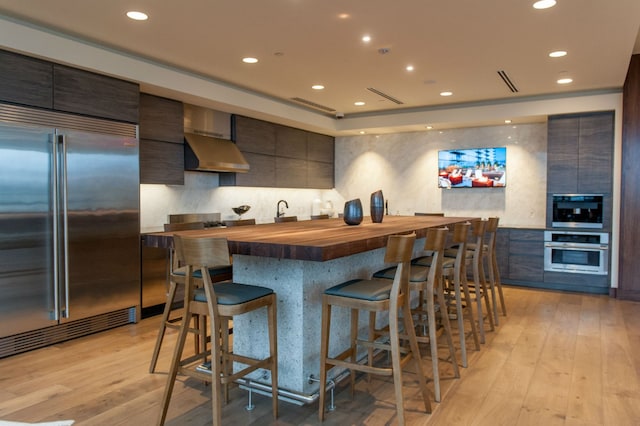 kitchen featuring stainless steel built in fridge, butcher block counters, light hardwood / wood-style floors, and wall chimney range hood