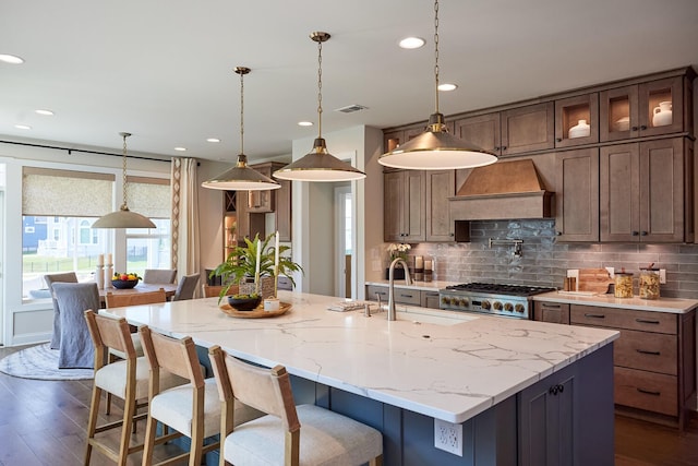 kitchen featuring premium range hood, hanging light fixtures, a kitchen island with sink, and dark hardwood / wood-style flooring