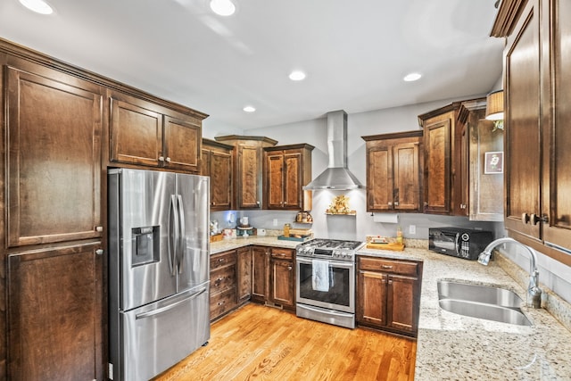 kitchen with light wood-type flooring, light stone counters, sink, wall chimney range hood, and appliances with stainless steel finishes