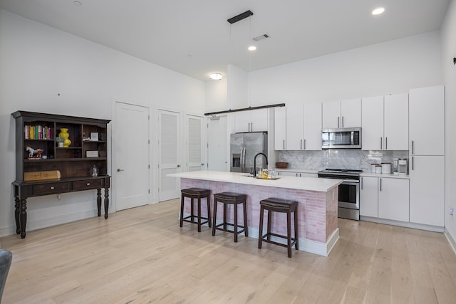 kitchen featuring white cabinets, decorative light fixtures, a center island with sink, appliances with stainless steel finishes, and light wood-type flooring