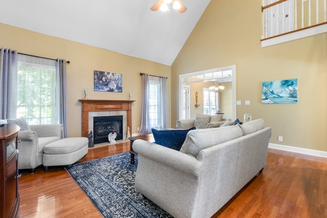 living room featuring hardwood / wood-style flooring, ceiling fan with notable chandelier, a tiled fireplace, and high vaulted ceiling