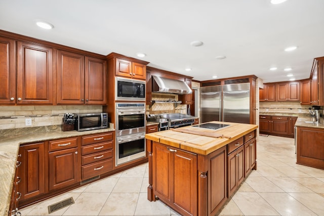 kitchen featuring light tile patterned floors, tasteful backsplash, wall chimney range hood, built in appliances, and butcher block counters