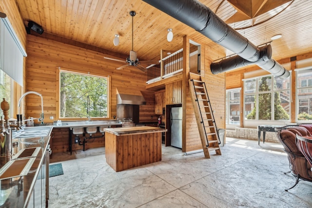 kitchen with a kitchen island, wooden walls, wall chimney exhaust hood, stainless steel refrigerator, and sink