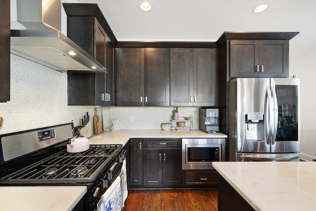 kitchen featuring wall chimney range hood, dark wood-type flooring, light stone counters, decorative backsplash, and appliances with stainless steel finishes