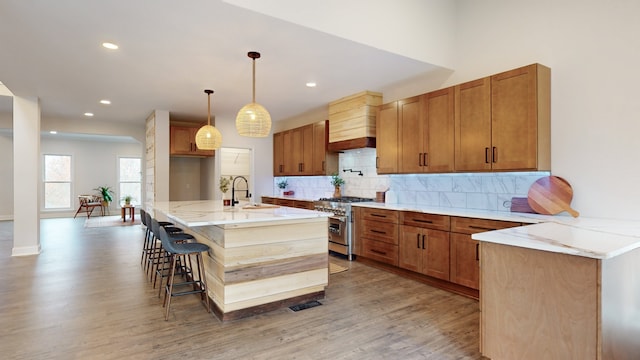 kitchen with a kitchen island with sink, light hardwood / wood-style flooring, decorative light fixtures, custom exhaust hood, and stainless steel range