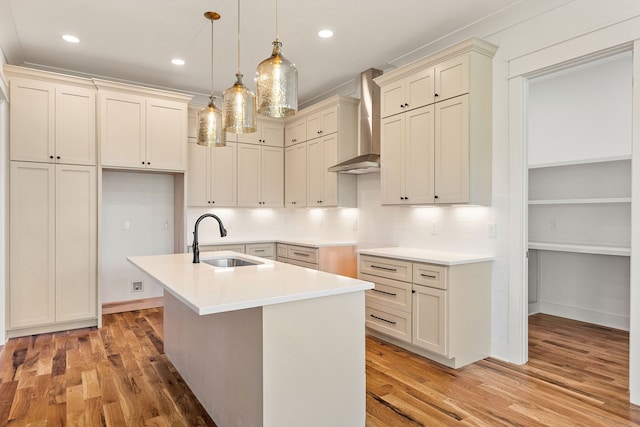 kitchen with an island with sink, sink, wall chimney range hood, light hardwood / wood-style flooring, and decorative light fixtures