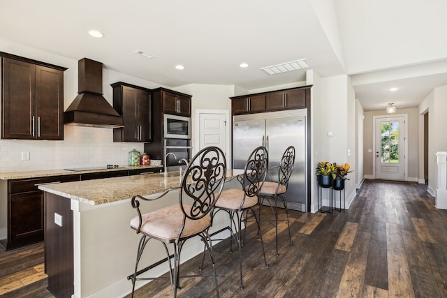 kitchen with built in appliances, an island with sink, premium range hood, light stone counters, and dark hardwood / wood-style flooring