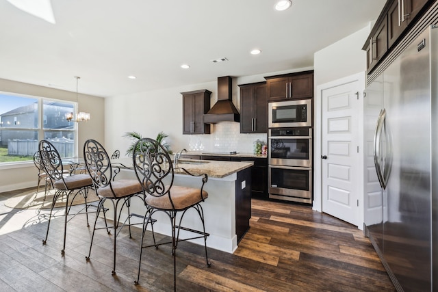 kitchen with a kitchen island with sink, dark hardwood / wood-style floors, built in appliances, hanging light fixtures, and custom exhaust hood