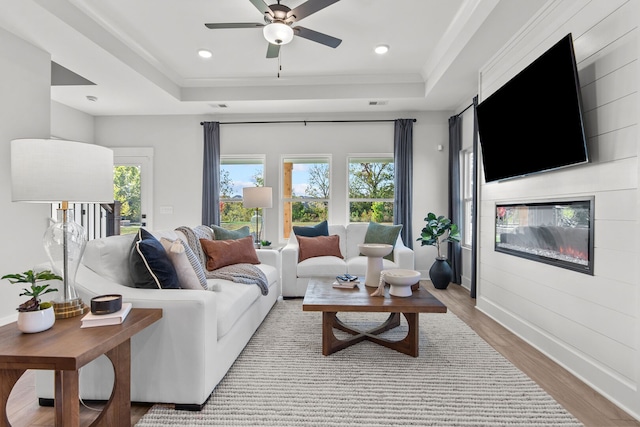 living room with ceiling fan, a tray ceiling, a fireplace, light wood-type flooring, and crown molding