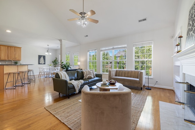 living room with ceiling fan with notable chandelier, high vaulted ceiling, ornate columns, and light hardwood / wood-style flooring