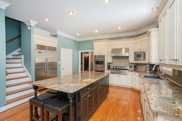 kitchen with a kitchen island, light stone counters, ventilation hood, sink, and built in appliances