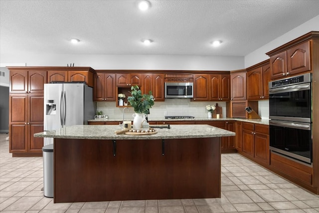 kitchen featuring a kitchen island with sink, light tile patterned floors, light stone countertops, appliances with stainless steel finishes, and tasteful backsplash