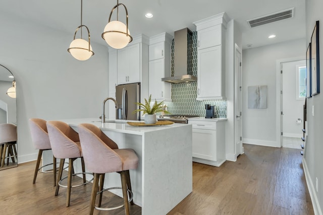 kitchen featuring wall chimney range hood, stainless steel fridge, pendant lighting, an island with sink, and white cabinetry