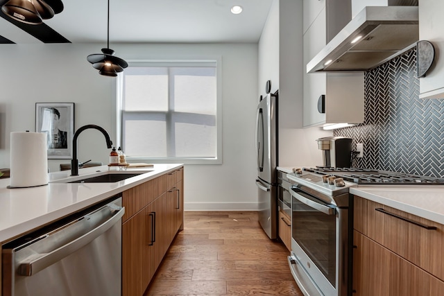 kitchen featuring stainless steel appliances, wall chimney exhaust hood, sink, light hardwood / wood-style flooring, and decorative light fixtures