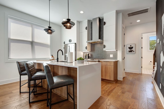 kitchen featuring wall chimney exhaust hood, a center island with sink, light hardwood / wood-style flooring, and white cabinetry