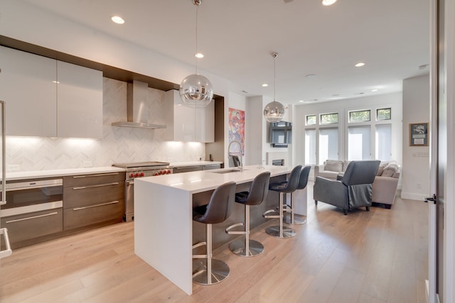 kitchen with wall chimney range hood, stainless steel stove, an island with sink, and light wood-type flooring