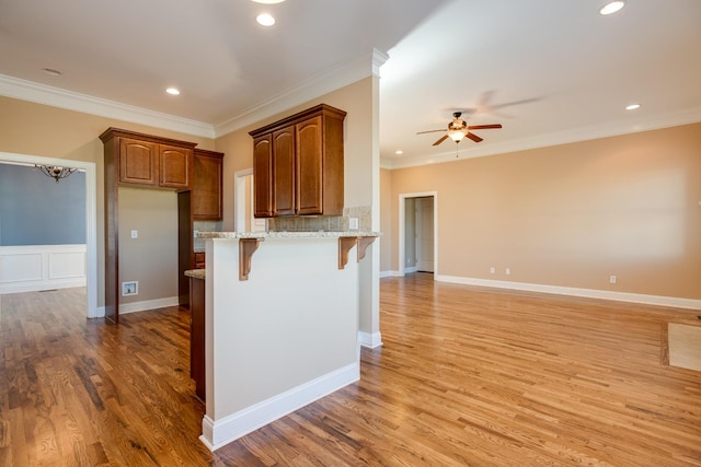 kitchen featuring light wood finished floors, light stone counters, a breakfast bar, ornamental molding, and ceiling fan with notable chandelier