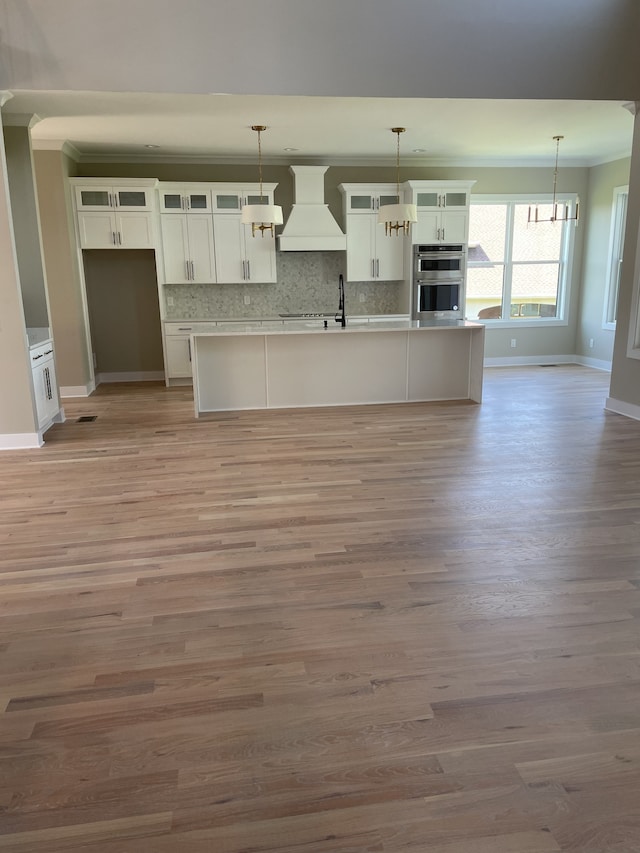 kitchen with light wood-type flooring, a kitchen island with sink, double oven, premium range hood, and pendant lighting