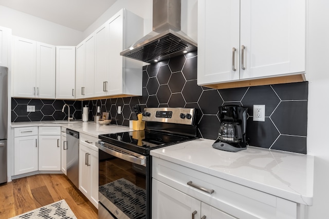 kitchen with tasteful backsplash, white cabinets, wall chimney range hood, light hardwood / wood-style flooring, and appliances with stainless steel finishes