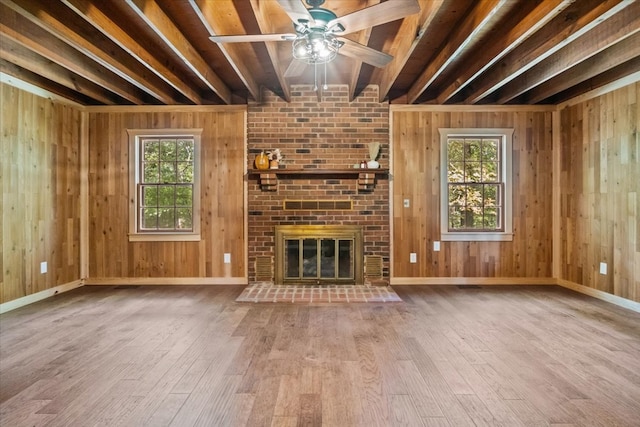 unfurnished living room with beam ceiling, light wood-type flooring, a brick fireplace, plenty of natural light, and wooden walls