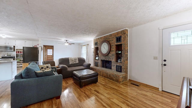 living room with a brick fireplace, a textured ceiling, light wood-type flooring, and ceiling fan