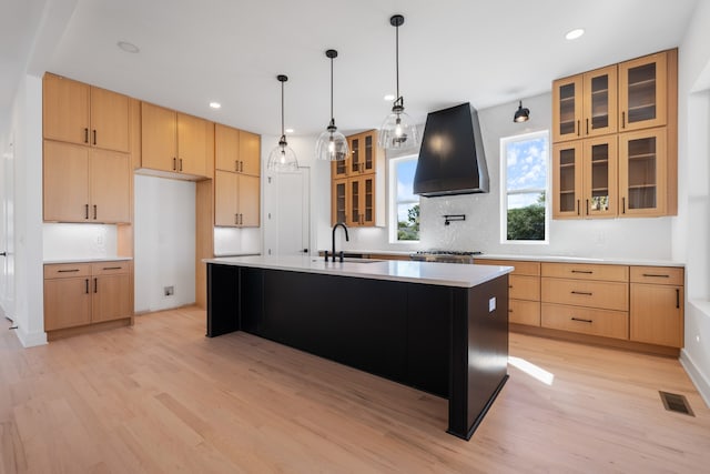 kitchen featuring a kitchen island with sink, wall chimney exhaust hood, sink, light wood-type flooring, and tasteful backsplash