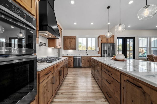 kitchen with custom exhaust hood, light stone counters, appliances with stainless steel finishes, light hardwood / wood-style flooring, and decorative light fixtures