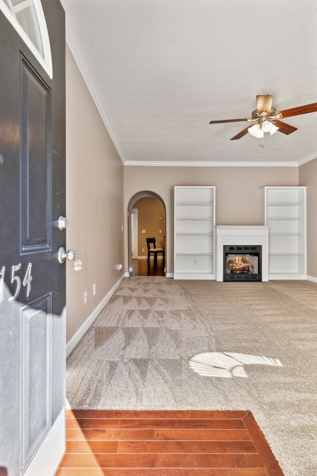 unfurnished living room with arched walkways, carpet flooring, baseboards, ornamental molding, and a glass covered fireplace