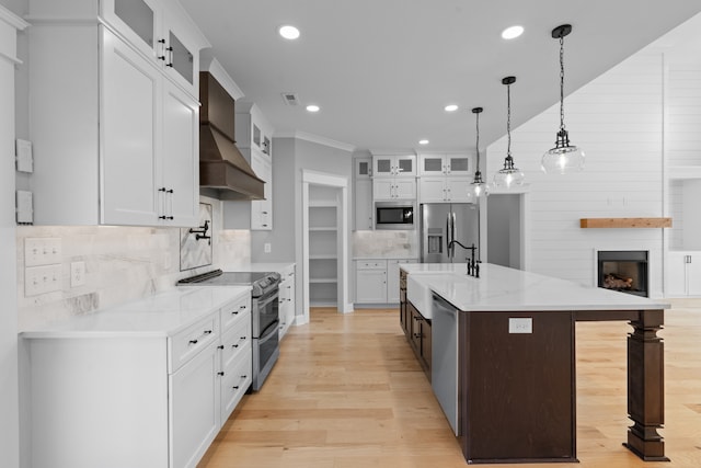 kitchen featuring a large island with sink, wall chimney range hood, white cabinets, decorative light fixtures, and appliances with stainless steel finishes