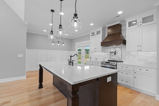 kitchen featuring stainless steel range, custom range hood, light hardwood / wood-style flooring, a center island with sink, and white cabinets