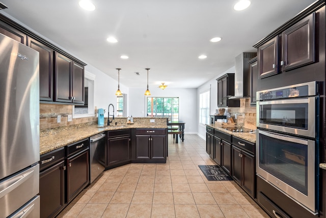 kitchen with wall chimney range hood, sink, backsplash, stainless steel appliances, and decorative light fixtures