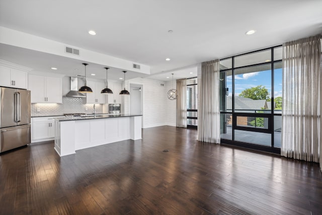 kitchen with wall chimney range hood, stainless steel fridge, hanging light fixtures, white cabinetry, and dark wood-type flooring