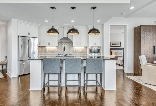 kitchen with appliances with stainless steel finishes, an island with sink, wall chimney exhaust hood, pendant lighting, and white cabinets