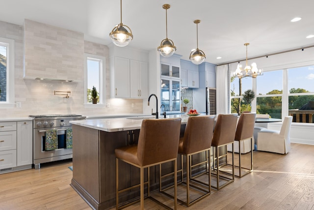 kitchen featuring decorative backsplash, high end stove, a center island with sink, white cabinetry, and hanging light fixtures