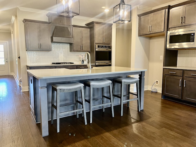 kitchen featuring hanging light fixtures, an island with sink, appliances with stainless steel finishes, custom exhaust hood, and ornamental molding