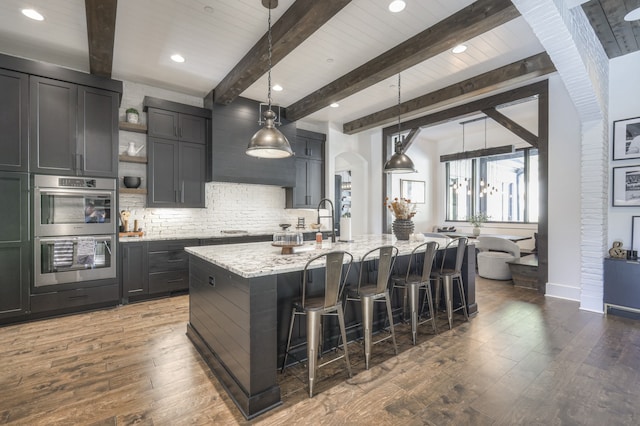 kitchen with light stone countertops, appliances with stainless steel finishes, hanging light fixtures, beam ceiling, and a center island with sink