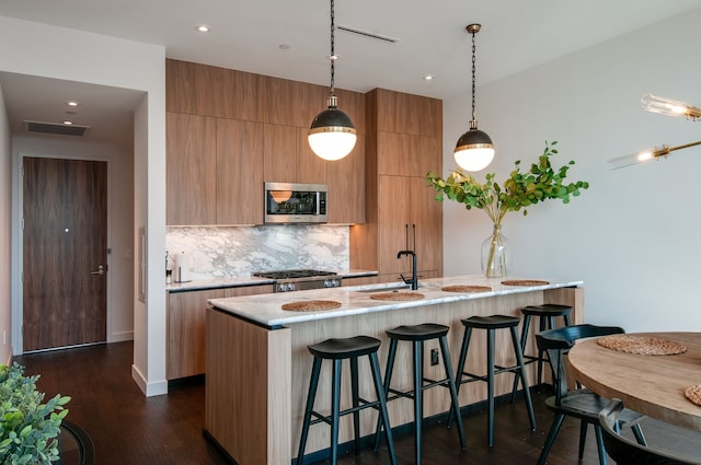 kitchen with appliances with stainless steel finishes, backsplash, dark hardwood / wood-style flooring, decorative light fixtures, and a kitchen island with sink