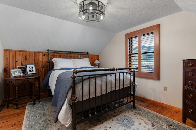 bedroom with light hardwood / wood-style floors, wood walls, a textured ceiling, and vaulted ceiling