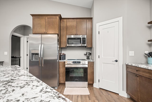 kitchen featuring light stone countertops, light wood finished floors, lofted ceiling, arched walkways, and stainless steel appliances