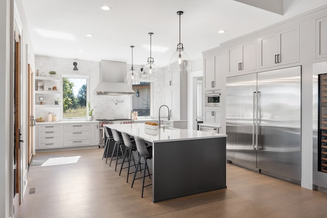 kitchen featuring white cabinets, a kitchen island with sink, pendant lighting, wall chimney exhaust hood, and high end appliances