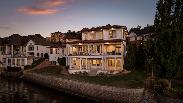 back house at dusk with a yard, a water view, and a balcony