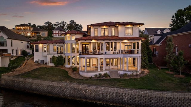 back house at dusk with a patio, a lawn, and a balcony