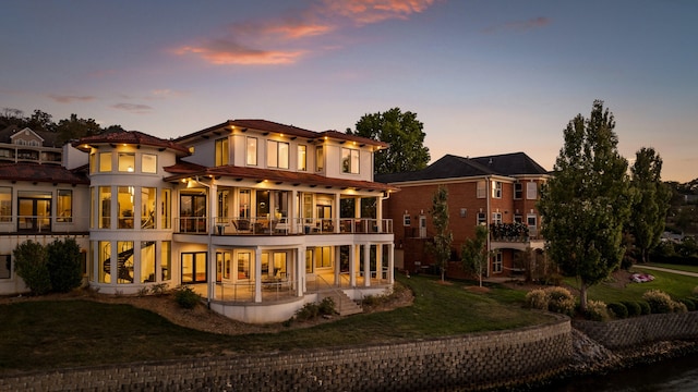 back house at dusk with a balcony, a patio area, and a lawn