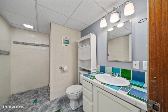 bathroom featuring a paneled ceiling, a shower, tile walls, toilet, and vanity