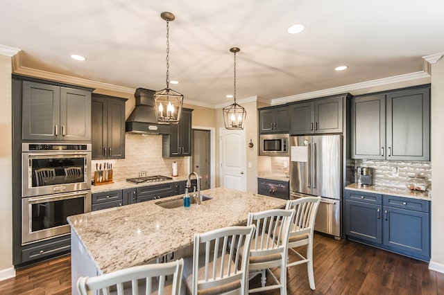 kitchen with sink, hanging light fixtures, stainless steel appliances, custom range hood, and a center island with sink